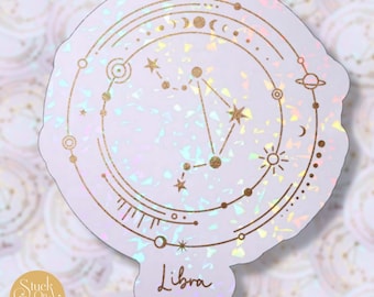 May include: A holographic sticker with a gold outline of the Libra constellation and the word "Libra" written below. The background is a light purple with a glittery texture.