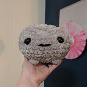 May include: A gray crocheted plushie with large black eyes and a small, stitched smile. The round, textured toy has small, stubby legs and is held in a hand.