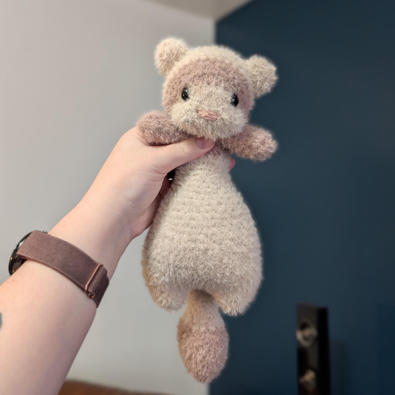 Op de afbeelding: Een handgemaakt fret speelgoed in beige en roze tinten. Het fret heeft een pluizige kop, kleine oren en een lang lichaam met een staart. Het speelgoed wordt in een hand gehouden, wat de zachte textuur en de gedetailleerde haakwerk benadrukt.