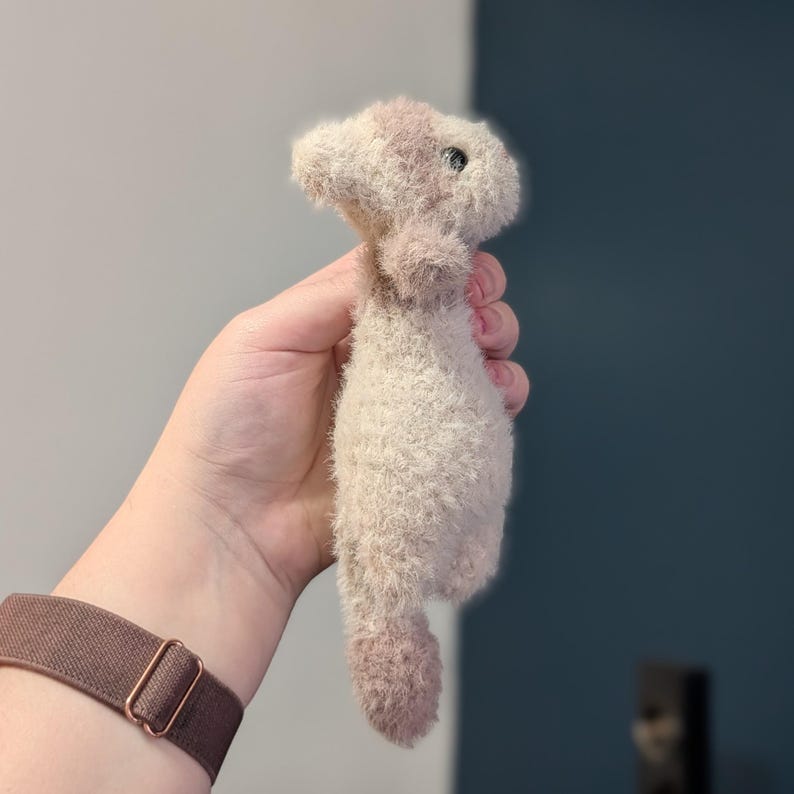 Op de afbeelding: Een handgemaakt pluche speelgoed in de vorm van een klein dier, met een cr&egrave;mekleurig lichaam en roze accenten op het hoofd en de voeten. Het speelgoed heeft een zachte textuur en een klein, donker oog. Het speelgoed wordt in een hand gehouden.