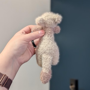 Op de afbeelding: Een klein, beige pluche speelgoed in de vorm van een poedel, vastgehouden in een hand. Het speelgoed heeft een zachte textuur en een roze staart en poten. De achtergrond is een mix van grijs en blauw.