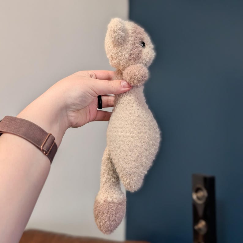 Op de afbeelding: Een handgemaakt pluche speelgoed in de vorm van een kat, met een zachte, getextureerde stof in tinten beige en lichtbruin. De kat heeft een afgerond lichaam, een klein hoofd met donkere ogen en een pluizige staart. Het speelgoed wordt door een hand vastgehouden.