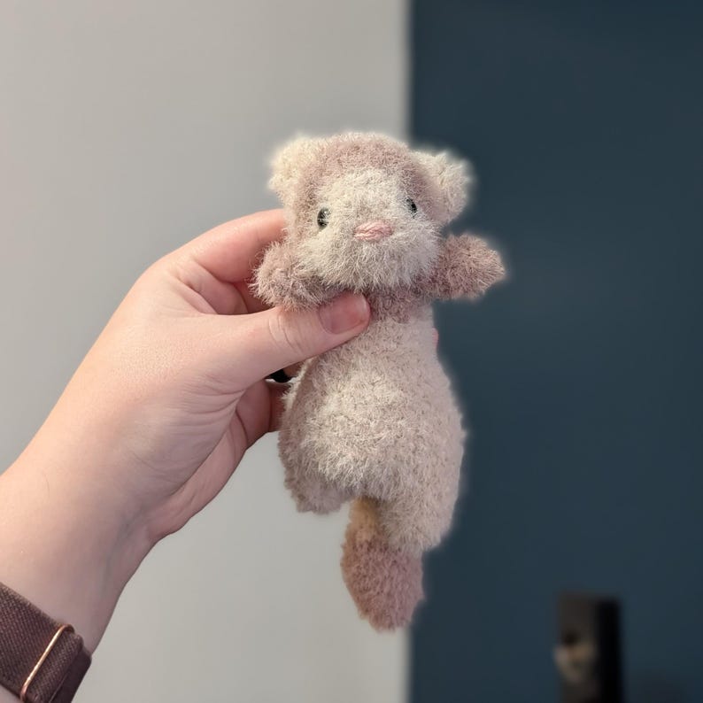Op de afbeelding: Een handgemaakte pluche kat in cr&egrave;me- en mauve-tinten. De kat heeft kleine zwarte ogen, een roze neus en een pluizige staart. Het speelgoed wordt in de hand van een persoon gehouden tegen een neutrale achtergrond.