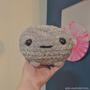 May include: A grey, plush, crocheted creature with large black eyes and a small, smiling mouth. The handmade toy has a round body and small feet. The background includes a white surface and a pink and white circular design.