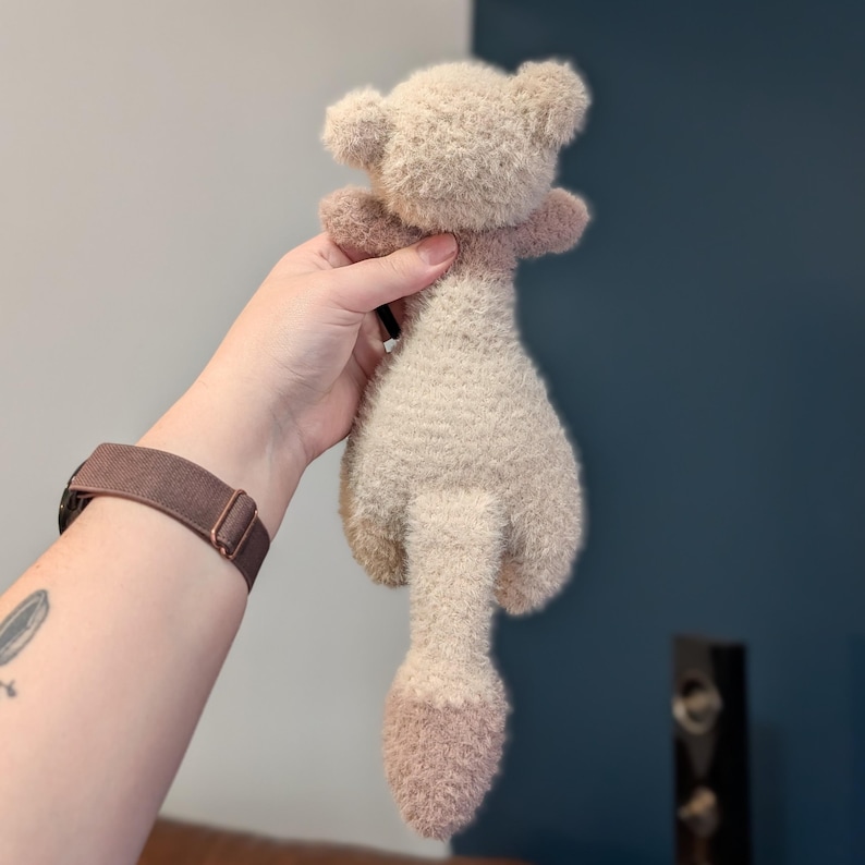 Op de afbeelding: Een handgemaakt pluche berenspeelgoed in een lichtbeige kleur met een donkerdere beige staart en poten. De beer heeft kleine oren en een zachte, fuzzy textuur. Het speelgoed wordt door een persoon omhoog gehouden.