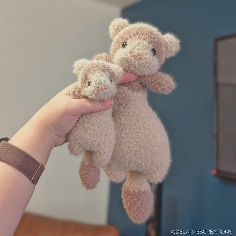 Op de afbeelding: Twee pluche teddyberen in een lichtbruin kleurenschema. De grotere beer wordt door een persoon vastgehouden, met de kleinere beer ervoor. De beren hebben zachte, getextureerde vacht, met donkerbruine accenten op de oren, poten en snuit.
