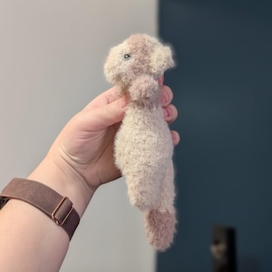 Op de afbeelding: Een klein, pluche speelgoed in beige en roze tinten, dat op een wezen lijkt. Het speelgoed heeft een zachte textuur en een afgeronde vorm, met een lichtere kop en een donkerroze staart. Het wordt tegen een neutrale achtergrond gehouden.