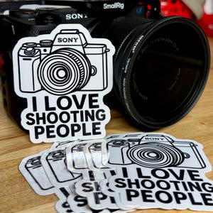 May include: A black Sony camera with a lens and several white stickers. The stickers feature a camera illustration and the text "I LOVE SHOOTING PEOPLE". The camera is on a wooden surface.