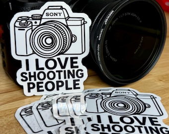 May include: A black Sony camera with a lens and several white stickers. The stickers feature a camera illustration and the text "I LOVE SHOOTING PEOPLE". The camera is on a wooden surface.