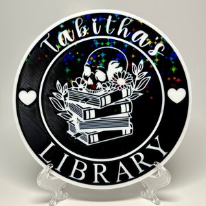 May include: A black and white round ceramic plate with a white skull and stack of books design. The plate has a glittery black background with white stars. The text "Tabitha's Library" is written in a white cursive font around the design.