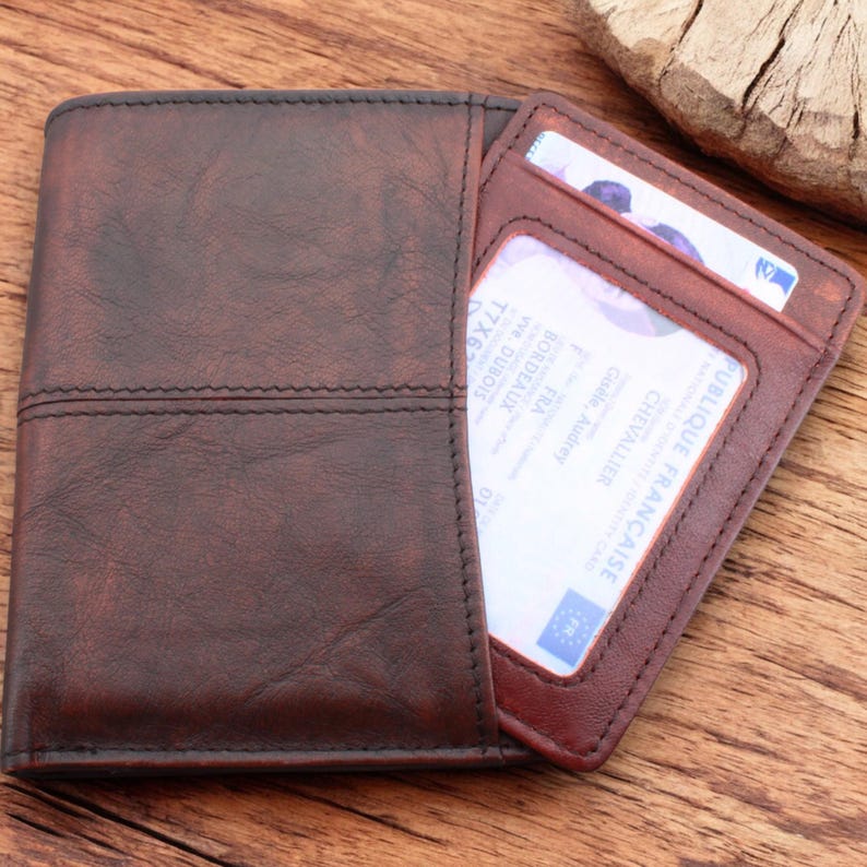May include: A brown leather wallet with a stitched design. The wallet has a flap that opens to reveal two card slots. The card slots are visible and contain cards with French text.