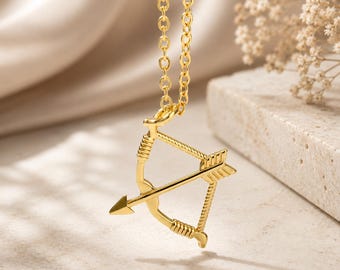 May include: A gold-toned necklace featuring a pendant shaped like a bow and arrow. The pendant has a polished finish with textured detailing on the bow limbs and the arrow shaft. The piece hangs from a delicate, matching gold-toned link chain.