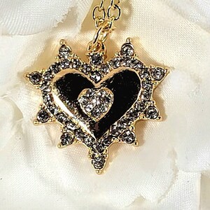 May include: A gold necklace with a black heart pendant surrounded by clear rhinestones. The heart has a smaller heart inside with clear rhinestones.