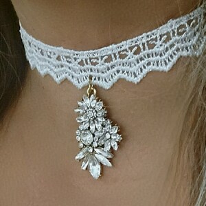 Może przedstawiać: Biała koronkowa chokerek z wiszącym kryształowym wisiorkiem. Wisiorek wykonany jest z przezroczystych cyrkonii i ma złote wykończenie.