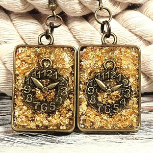May include: Gold leaf earrings with a clock face design. The earrings are made of bronze and have a square shape. The clock face is surrounded by gold leaf and has Roman numerals.