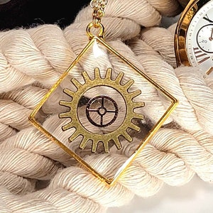 May include: A gold necklace with a square pendant featuring a gold gear design inside a clear resin casing. The necklace is displayed on a white rope.