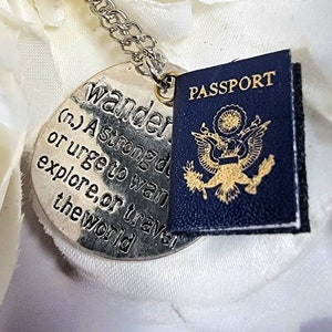 以下が含まれることがあります： 「wander (n.) A strong desire or urge to want to explore, or travel the world」と刻まれた丸いチャームが付いたシルバーネックレス。小さな青い布製の旅券がチャームに付けられています。