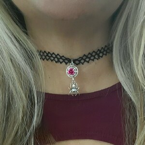 May include: A black choker necklace with a silver spider charm and a red gem. The spider is hanging from a web-shaped pendant.