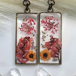 Puede incluir: Un par de pendientes rectangulares de resina con flores secas y escamas doradas. Los pendientes son de color marrón claro con un gancho plateado.