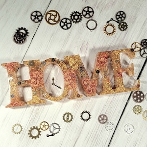 May include: A gold and copper colored resin "HOME" sign with a clock face and gears embedded in the resin. The sign is surrounded by small metal gears on a white wooden surface.