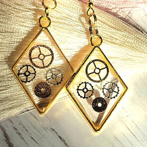 May include: Gold diamond-shaped earrings with clear resin and black metal gears inside.