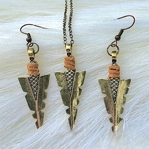 Könnte beinhalten: Ein Set aus goldfarbenen Pfeilspitzen-Ohrringen und einer Halskette. Die Pfeilspitzen sind mit einem strukturierten Muster verziert und haben oben einen braunen Lederüberzug.