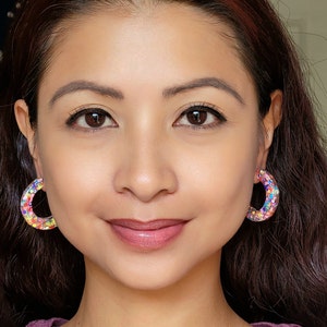 May include: A woman with long brown hair wearing a purple shirt and colorful hoop earrings.