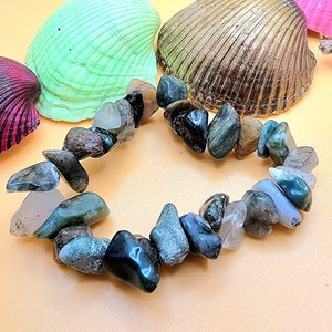 Puede incluir: Un brazalete de piedras preciosas verdes, marrones y transparentes con piedras de forma irregular. El brazalete está sobre un fondo naranja claro con conchas.