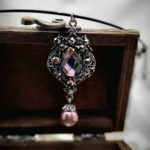 Op de afbeelding: Een zilveren ketting met een hanger met een grote, gefacetteerde, paarse edelsteen omringd door kleinere zwarte edelstenen. De hanger is bevestigd aan een zwart koord en hangt aan een houten doos.