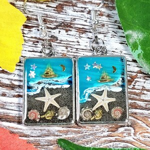 May include: A pair of silver earrings with a blue, white, and gold seascape design. The earrings feature a seashell, a starfish, and a crescent moon.