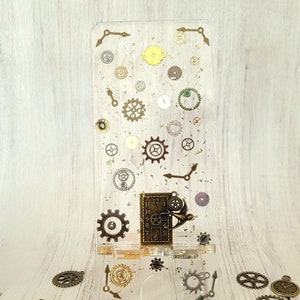 May include: A clear phone stand with a clock face and gears embedded in the resin. The stand is surrounded by loose gears and clock hands.