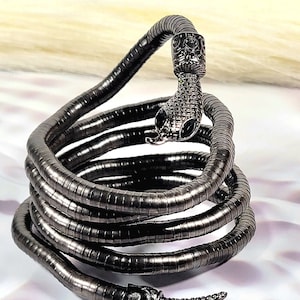 May include: A black metal snake-shaped bracelet with a coiled design. The bracelet is made of multiple metal rings that are connected together to form a snake-like shape. The head of the snake is detailed with a textured design and black gemstones for eyes.