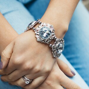 Puede incluir: Pulsera plateada con grandes piedras preciosas ovaladas de color azul claro. La pulsera tiene un diseño ornamental de filigrana. También se ve un anillo plateado con una gema grande y transparente y un anillo de flores plateado.
