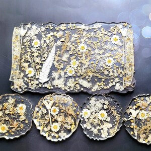 May include: A set of five resin coasters featuring pressed dried flowers and leaves in shades of white and gold. The coasters are in a rectangular shape with a wavy edge.