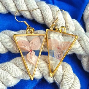 K&ouml;nnte beinhalten: Ein Paar goldene Dreiecksohrringe mit zarten gepressten rosa Bl&uuml;ten, die in klarem Harz eingeschlossen sind.