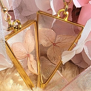 K&ouml;nnte beinhalten: Ein Paar goldene Dreiecksohrringe mit zarten gepressten rosa Bl&uuml;ten, die in klarem Harz eingeschlossen sind.