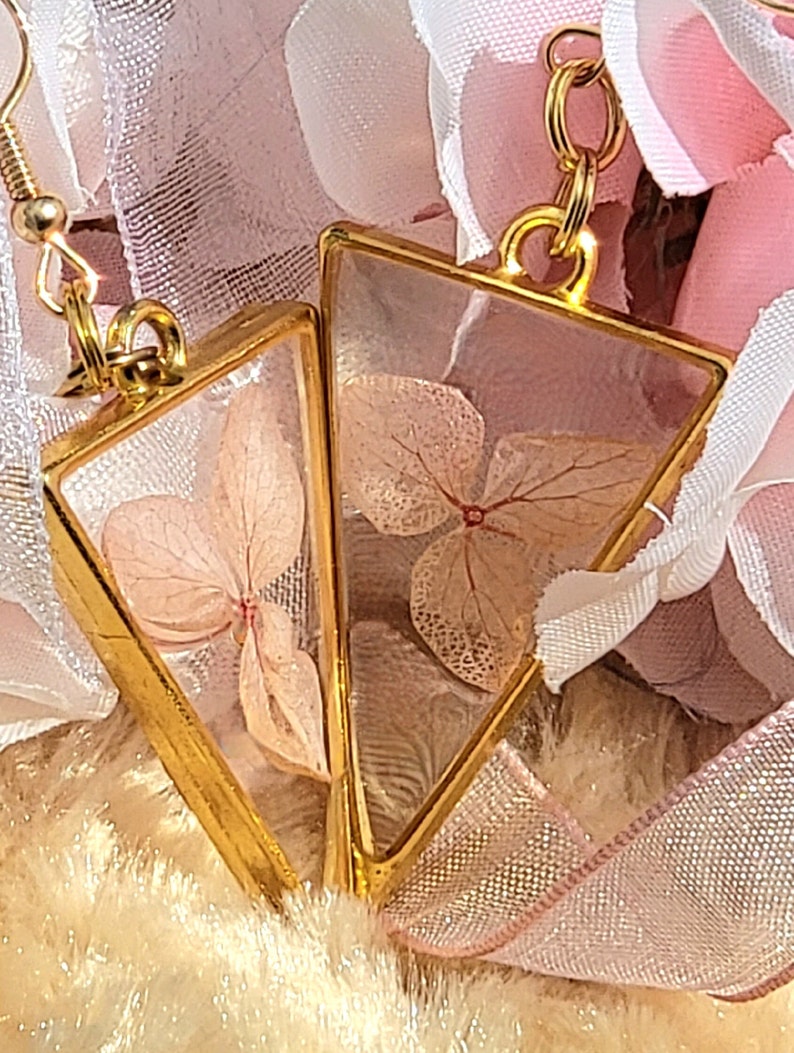 K&ouml;nnte beinhalten: Ein Paar goldene Dreiecksohrringe mit zarten gepressten rosa Bl&uuml;ten, die in klarem Harz eingeschlossen sind.