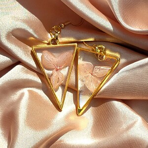 K&ouml;nnte beinhalten: Ein Paar goldene Dreiecksohrringe mit zarten, gepressten rosa Bl&uuml;ten im Inneren. Die Ohrringe liegen auf einem hellrosa Satin-Hintergrund.