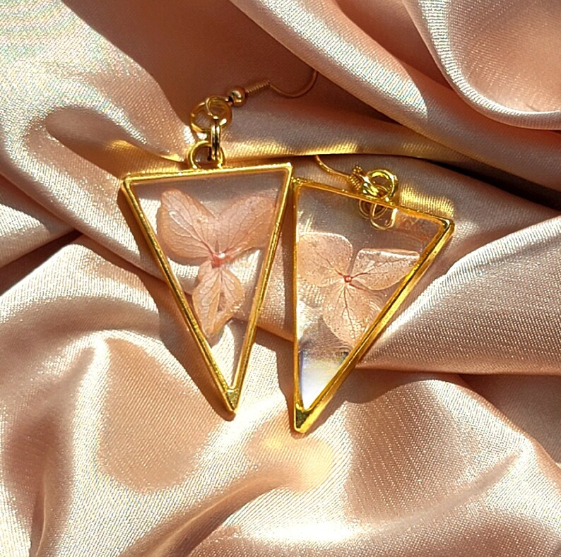 K&ouml;nnte beinhalten: Ein Paar goldene Dreiecksohrringe mit zarten, gepressten rosa Bl&uuml;ten im Inneren. Die Ohrringe liegen auf einem hellrosa Satin-Hintergrund.