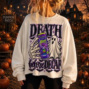 Op de afbeelding: Een crèmekleurig sweatshirt met lange mouwen en een grafische print van een skeletthand die een paars Alani energiedrankblikje vasthoudt naast een wit spook. De tekst luidt DEATH before DECAF in dikke paarse letters met kleine zwarte vleermuizen.