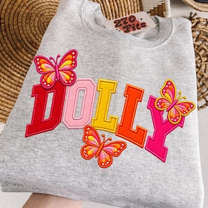 May include: A heather gray crewneck sweatshirt featuring the word DOLLY in large, multi-colored block letters. The design includes three embroidered butterflies in shades of pink, orange, and yellow. A tag labeled 270 Fits is visible at the neck.
