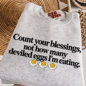 Peut inclure: Un sweat-shirt à col rond gris clair avec le texte noir, Count your blessings, not how many deviled eggs I'm eating. Sous le texte se trouvent trois illustrations d'œufs farcis coupés en deux avec des jaunes jaunes et des blancs blancs.