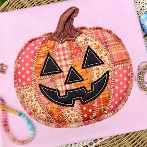 May include: A fabric pumpkin appliqué featuring a patchwork design of various orange, pink, and white patterned textiles. The pumpkin has a brown stem and black felt facial features, including triangular eyes and a jagged mouth, all secured with visible zigzag stitching.