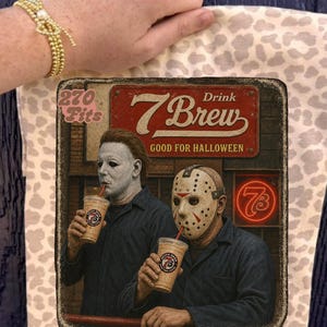 Pode incluir: Uma bolsa tote bege com estampa de leopardo marrom claro apresentando um gráfico de dois personagens de filmes de terror bebendo de copos de café 7 Brew. O texto diz 270 Fits, Drink 7 Brew e GOOD FOR HALLOWEEN com um letreiro neon.