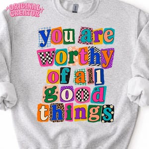 May include: A light gray crewneck sweatshirt featuring the text "YOU ARE WORTHY OF ALL GOOD THINGS" in a multi-colored, collage-style font. Each letter is enclosed in a uniquely patterned, colorful rectangular patch with various prints.