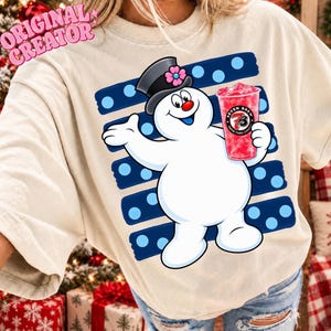 Puede incluir: Una camiseta beige con el gráfico de un muñeco de nieve blanco que lleva un sombrero de copa negro con una flor rosa. El muñeco sostiene un vaso rojo con el logo de Seven Brew. Detrás hay rayas azul oscuro con puntos azul claro.