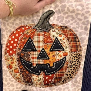May include: A fabric pumpkin decoration featuring a patchwork design of various autumn-themed patterns including orange polka dots, plaid, floral prints, and leopard print. It has a black stitched jack-o'-lantern face and a dark green fabric stem.
