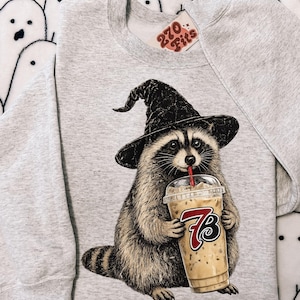 Pode incluir: Uma camisola de gola redonda cinzenta clara com o gráfico de um guaxinim a usar um chapéu de bruxa preto e a beber café gelado de um copo de plástico transparente com uma palhinha vermelha. O copo apresenta o texto 7B em vermelho e preto.