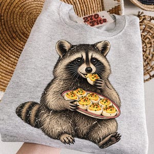 May include: A light gray crewneck sweatshirt featuring a graphic illustration of a raccoon holding a platter of deviled eggs and eating one. The inner neck label displays the text 270 Fits in red lettering on a white background.