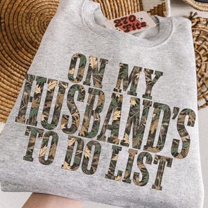 May include: A light gray crewneck sweatshirt featuring the text ON MY HUSBAND'S TO DO LIST in a camouflage leaf pattern. The garment includes a small white tag near the neckline with the text 270 Fits printed in red and black lettering.